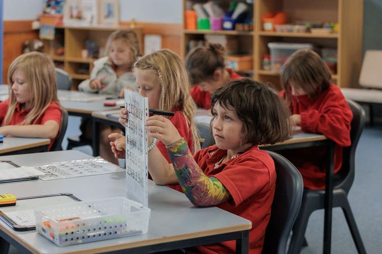 Image of students seated at tables in a classroom. One girl is holding up her writing sheet in front of her.
