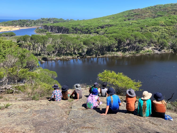 Students seated on a large flat rock overlooking a lagoon surrounded by bush.